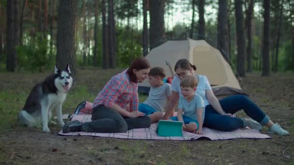 Two 30s mothers and two sons watching tablet computer laying on picnic blanket on summer camping vacation with tent in forest. Happy family with Siberian Husky dog. Slow motion, steadicam shot. — Stock Video
