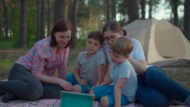 Duas mães dos anos 30 e dois filhos assistindo tablet computador deitado em cobertor de piquenique no verão acampando férias com barraca na floresta. Família feliz com cão Husky siberiano. Movimento lento, steadicam tiro . — Vídeo de Stock