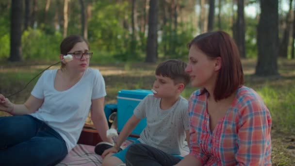 Dos madres y su hijo comiendo malvaviscos en palos. Dos mujeres y dos niños teniendo vacaciones de campamento de verano en el bosque. cámara lenta, disparo steadicam . — Vídeo de stock