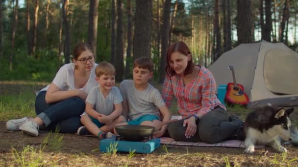 Happy family of two moms and two sons cooking on frying pan sitting on picnic blanket with Siberian Husky dog on summer camping vacation with tent in forest. Slow motion, steadicam shot. — Stock Video