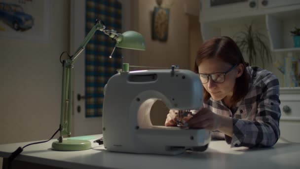 Jovem mulher de 30 anos ajustando máquina de costura que trabalha em casa. Costureira feminina em óculos costura sentado na sala de estar em casa . — Vídeo de Stock
