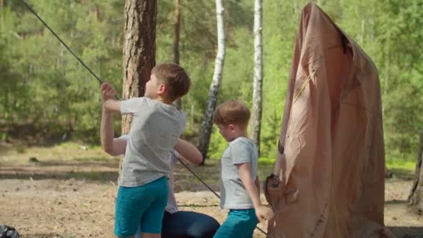 Dwóch chłopców pomaga dwóm mamom rozbić namiot w lesie dla lokalnej rodzinnej turystyki kempingowej. Rodzinna ekologiczna turystyka letnia. Zwolnij, Steadicam shot. — Wideo stockowe