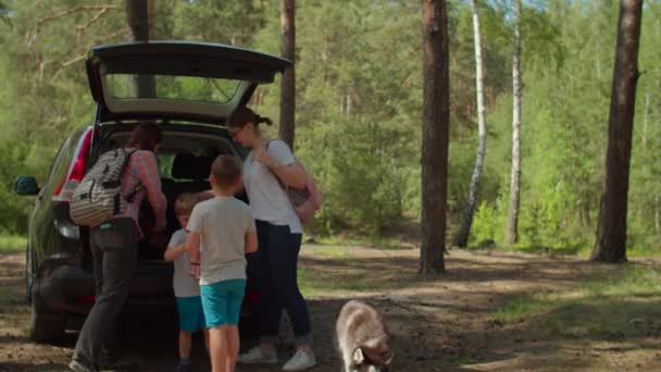 Two women, two boys having and Siberian Husky dog on summer camping vacation. Happy family of two mothers and two sons taking bags from car trunk and walking in forest. Slow motion, steadicam shot. — Stock Video