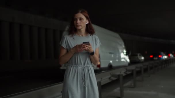 若い30代のブルネットの女性は、携帯電話で地図を検索しようとして、長い灰色のドレスで道に迷った。雨の日に暗い都市の車のトンネルの歩道に滞在女性. — ストック動画