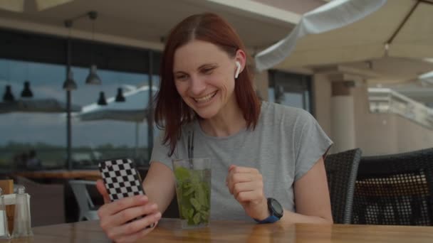 Mujer joven de 30 años hablando con un amigo por videollamada en el teléfono móvil y bebiendo bebida fresca con paja sentada sola en la cafetería de verano. Señora bebe mojito cóctel en la terraza usando el teléfono celular para charlar . — Vídeos de Stock