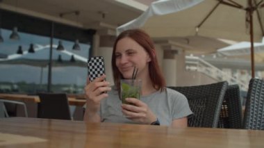 30 'lu yaşlardaki genç bir kadın cep telefonuyla arkadaşlarıyla konuşuyor ve yazlık kafede tek başına oturmuş pipetle serin içecekler içiyor. Bayan terasta mojito kokteyli içiyor cep telefonuyla sohbet ediyor..