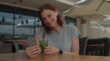 30 'lu yaşlardaki genç bir kadın cep telefonuyla arkadaşlarıyla konuşuyor ve yazlık kafede tek başına oturmuş pipetle serin içecekler içiyor. Bayan terasta mojito kokteyli içiyor cep telefonuyla sohbet ediyor..