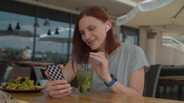携帯電話でビデオ通話で友人と話す若い30代の女性と夏のカフェで一人で座ってわらでクールな飲料を飲んでいます。女性は、チャットに携帯電話を使用してテラスでモヒートカクテルを飲む. — ストック動画