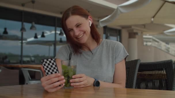 30多岁的女性通过电话视频和朋友聊天，夏天一个人坐在咖啡店里喝清凉的饮料。女士在阳台上喝莫吉托鸡尾酒，用手机聊天. — 图库视频影像