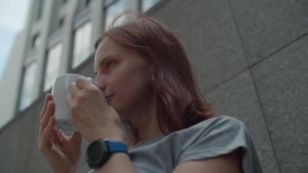 Jeune femme brune des années 30 buvant une tasse de café dans un café urbain. Dame dans les écouteurs sans fil aime boire des boissons chaudes au centre-ville. — Video