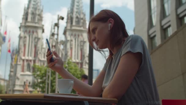 Młoda kobieta po trzydziestce w słuchawkach bezprzewodowych rozmawia z przyjacielem przez telefon komórkowy, siedzi w miejskiej kawiarni i pije kawę.. — Wideo stockowe