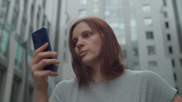 Joven mujer de negocios de 30 años hablando por videollamada en el teléfono móvil de pie cerca del centro de negocios en el centro. Mujer gerente tiene conversación en línea por celda al aire libre. De cerca. . — Vídeo de stock