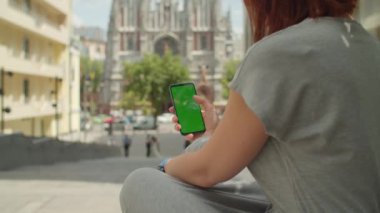 Şehir caddesinde oturan yeşil ekranlı cep telefonu kullanan genç bir kadın. Omuz manzaralı. Sokakta krom anahtarlı bir kadın cep telefonunu kaydırıyor..