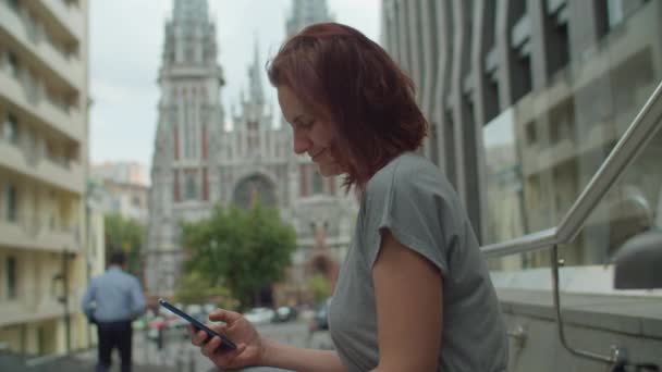 Молода 30 років жінка в бездротових навушниках, використовуючи мобільний телефон, що сидить на вулиці. Леді серфінг онлайн на мобільному телефоні на вулиці . — стокове відео