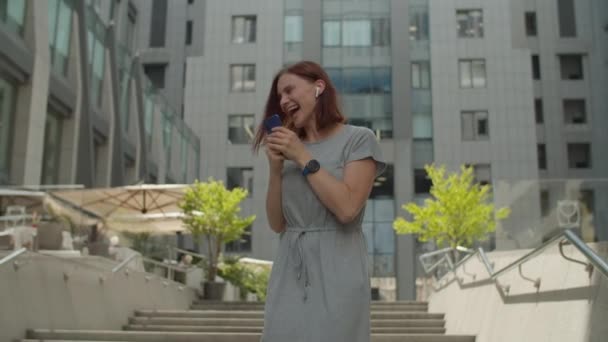 Jonge dertiger jaren zakenvrouw is erg blij om goed nieuws te lezen op mobiele telefoon staan in de buurt van business center in het centrum. Vrouw springen en dansen gelukkig op straat. — Stockvideo