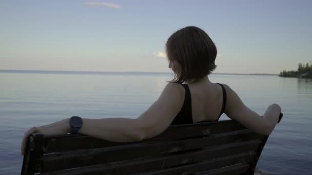 Young 30s woman in black dress sitting on bench on the riverside at the sunset. Lady enjoying her time alone by the evening pond. — Stock Video