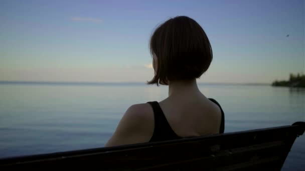 Młoda kobieta po trzydziestce w czarnej sukience, siedząca na ławce nad brzegiem rzeki o zachodzie słońca. Pani ciesząca się samotnością nad stawem.. — Wideo stockowe