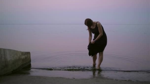 Jonge dertiger jaren vrouw in zwarte jurk die bij zonsondergang in het water staat. Barefoot dame knijpen een natte jurk in de avond vijver. — Stockvideo