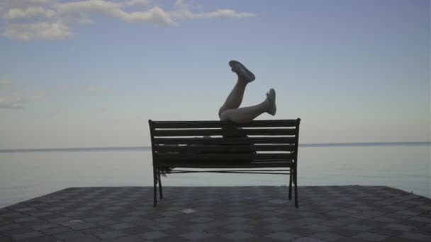 Giovane donna anni '30 in abito nero posa con le gambe alzate sulla panchina sul lungofiume al tramonto. Signora godendo il suo tempo da sola dallo stagno di sera. — Video Stock