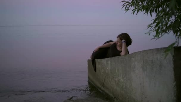 Eine junge Frau in schwarzem Kleid liegt bei Sonnenuntergang auf einer Brüstung am Wasser. Barfußdame genießt Entspannung allein am Abendweiher. — Stockvideo
