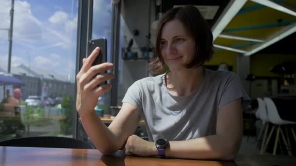 Νεαρή 30άρα γυναίκα μιλάει με βιντεοκλήση στο κινητό και χαμογελάει. Η κυρία μιλάει στο κινητό με ένα φίλο που κάθεται στο cafe της πόλης. — Αρχείο Βίντεο