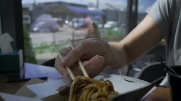 Gros plan de la main avec des baguettes mélangeant nouilles et viande et légumes dans une boîte en papier. asiatique boîte nourriture avec baguettes. — Video