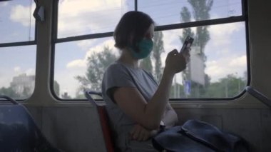 Yüz koruyucu maskeli genç bir kadın toplu taşıma aracına biniyor. Yüz maskeli kadın boş bir trende pencere kenarında cep telefonu kullanıyor..