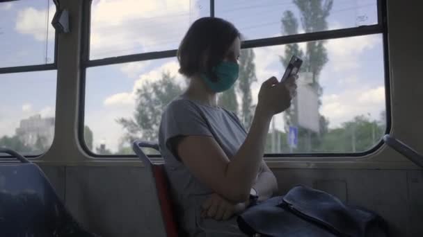 얼굴 보호 마스크를 쓴 젊은 여성 이 대중 교통에서 타고 있습니다. 창문 옆에 앉아 있는 휴대 전화를 사용하여 얼굴을 가리고 있는 여자. — 비디오