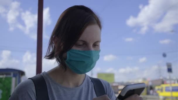 Ung kvinna i skyddande ansiktsmask med mobiltelefon i väntan på kollektivtrafik på stationen. Stadståg som kör på gatan. Kvinna med cell på transportstationen. — Stockvideo