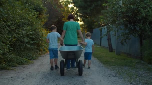 Familia de madre soltera y dos niños caminando por la calle del pueblo sosteniendo vagón al atardecer. cámara lenta, disparo steadicam . — Vídeos de Stock