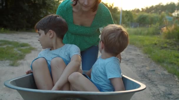 Een vrouw die een wagen duwt met kinderen erin. Alleenstaande moeder en twee zoons met trolley op dorpsstraat bij zonsondergang. Langzame beweging, standvastig schot.. — Stockvideo