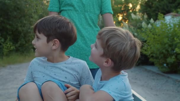 Familie van alleenstaande moeder en twee kinderen op dorpsstraat met wagen. Broers en zussen spelen zittend in de wagen. Vrouw trekt wagen met kinderen buiten. Langzame beweging, standvastig schot.. — Stockvideo