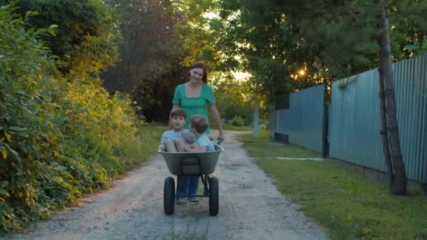 Een vrouw die een wagen duwt met kinderen erin. Alleenstaande moeder en twee zoons met trolley op dorpsstraat bij zonsondergang. Langzame beweging, standvastig schot.. — Stockvideo