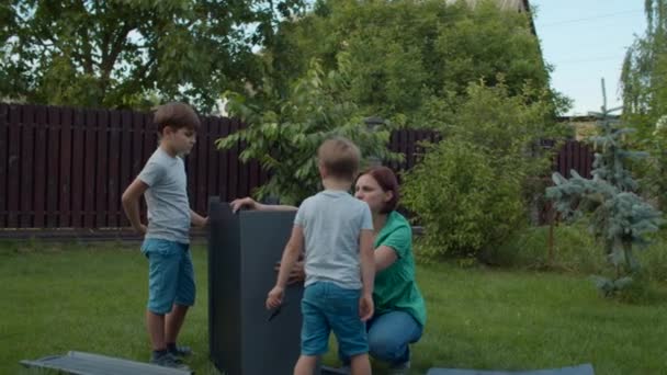 Two brothers helping young single mother to assemble furniture with instruction on backyard on sunny day. Happy family activities outdoors. Slow motion, steadicam shot. — Stock Video