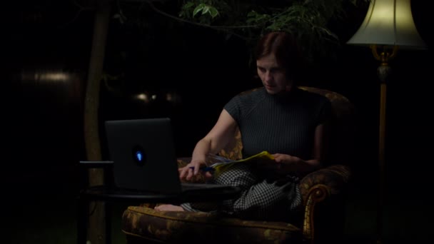 Młoda kobieta po trzydziestce studiuje online późno w nocy siedzi w przytulnym fotelu z lampą podłogową na podwórku. Dorosły student piszący w notatniku patrzący na laptopa. — Wideo stockowe