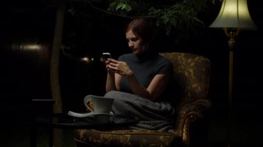 Genç mutlu kadın cep telefonundan iyi haberler okuyor ve gece geç saatlerde arka bahçesinde lamba olan rahat koltukta tek başına ellerini çırpıyor..