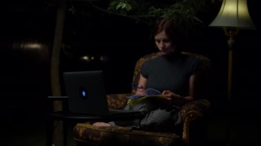30 'lu yaşlardaki genç bir kadın gece geç saatlere kadar online çalışmaktan rahat koltukta oturmaktan ve arka bahçesinde lamba taşımaktan yoruldu. Yetişkin öğrenci dizüstü bilgisayarını atıyor ve kapatıyor..