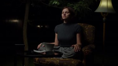 30 'lu yaşlardaki genç bir kadın meditasyona başlıyor geceleri rahat koltukta oturuyor. Meditasyon yaparken nefes alıp veren bir kadın..