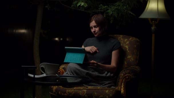 Young happy woman talks to friend by video call on tablet computer late at night sitting alone in cozy armchair with floor lamp in the backyard. — Stock Video