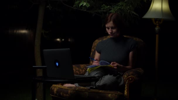 若い30代の女性が夜遅くに居心地の良いアームチェアに座って裏庭に床灯をオンラインで勉強するのに疲れている。ノートブックを捨ててノートパソコンを閉じる大人の学生. — ストック動画