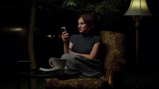 Jovem mulher feliz fala com amigo por videochamada no celular tarde da noite sentado sozinho em poltrona aconchegante com lâmpada de assoalho no quintal . — Vídeo de Stock