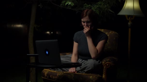 Young 30s woman watches scary movie on laptop late at night sitting in cozy armchair with floor lamp in the backyard. Woman empathizes with film heroes on laptop screen. — Stock Video