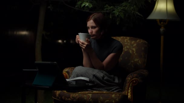 Młoda kobieta po trzydziestce ogląda zabawny film na tablecie późno w nocy siedząc w przytulnym fotelu z lampą podłogową na podwórku. Kobieta śmieje się patrząc na ekran gadżetu. — Wideo stockowe