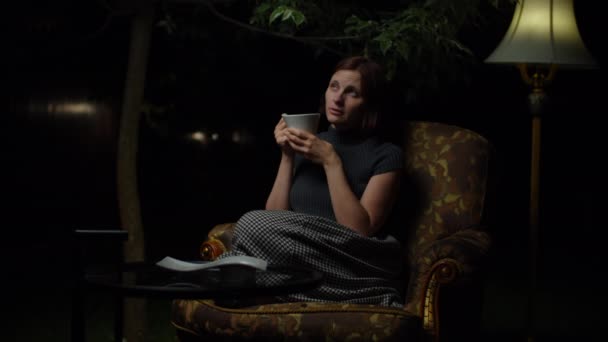 Jonge dertiger jaren vrouw denken tijdens het drinken van thee zitten in gezellige fauteuil met vloerlamp laat in de nacht in de achtertuin. — Stockvideo