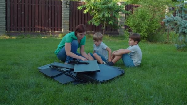 Rodzina młodej samotnej matki i dwójki dzieci montuje meble z instrukcją na podwórku w słoneczny dzień. Wesołych rodzinnych zajęć na świeżym powietrzu. Zwolnij, Steadicam shot. — Wideo stockowe