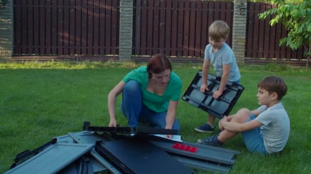 Two brothers helping young single mother to assemble furniture with instruction in backyard on sunny day. Happy family activities outdoors. Slow motion, steadicam shot. — Stock Video