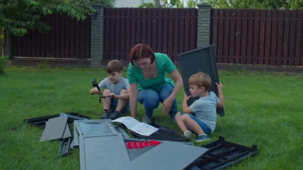 Два брата помогают молодой матери-одиночке собирать мебель с инструкциями на заднем дворе в солнечный день. Счастливая семейная жизнь на свежем воздухе. Медленное движение, устойчивый снимок . — стоковое видео