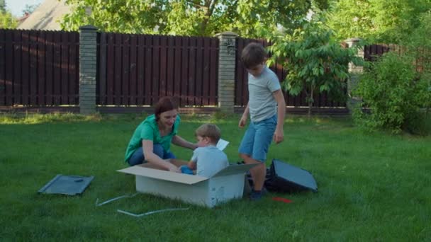 Mutter und zwei Kinder amüsieren sich an sonnigen Tagen mit einer großen Kiste im Hinterhof. Glückliche Familie spielt mit Lieferkarton auf dem Rasen. Zeitlupe, Steadicam-Aufnahme. — Stockvideo