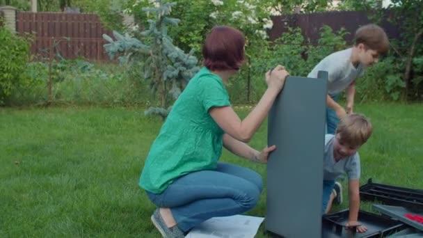 Twee broers helpen jonge alleenstaande moeder om meubels te monteren met instructie in de achtertuin op zonnige dag. Gelukkige familie activiteiten buiten. Langzame beweging, standvastig schot.. — Stockvideo