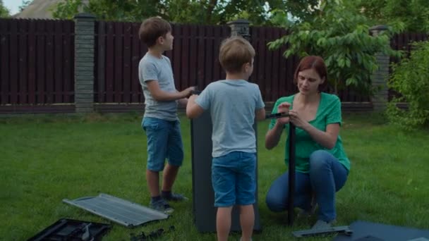Family of young single mother and two kids assembles furniture with instruction in backyard on sunny day. Happy family weekend activities outdoors. Slow motion, steadicam shot. — Stock Video